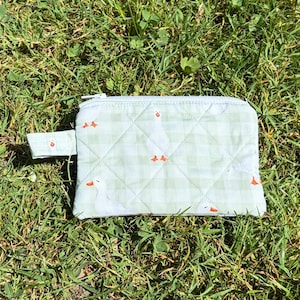 May include: A rectangular quilted zipper pouch featuring a light green and white gingham check pattern with a repeating print of white geese with orange beaks and feet. The pouch includes a small matching fabric tab on the left side.