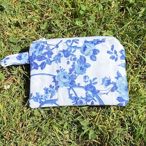 May include: A rectangular fabric zipper pouch featuring a white background with a repeating blue floral pattern of roses and leaves. The pouch includes a matching fabric wrist strap attached to the side and a white zipper closure along the top edge.
