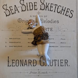 Könnte beinhalten: Vintage Porzellanfigur einer Frau im Badeanzug, mit rotem Stirnband, auf einem Notenpapierhintergrund. Das Notenblatt trägt die Aufschrift "Sea-Side Sketches" und listet verschiedene Orte auf. Die Figur ist etwa 10 cm groß.
