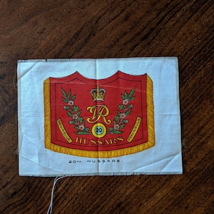Könnte beinhalten: Ein weißer Stoffabzeichen mit einem roten und goldenen Wappen, das eine Krone und den Buchstaben "R" mit der Zahl "20" darunter zeigt. Das Wappen ist von einem Kranz aus grünen Blättern und rosa Blüten umgeben. Der Text "HUSSARS" befindet sich unter dem Wappen, und der Text "20th. HUSSARS." darunter.