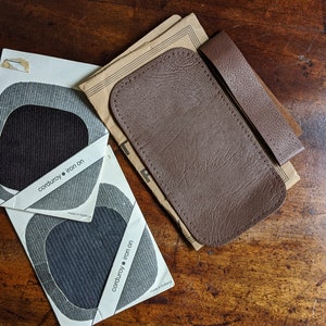 May include: A collection of craft materials displayed on a wooden surface. The image shows two corduroy iron-on patches in grey and black, alongside brown leather pieces. The patches have a grey and white design, and the leather pieces are various shapes.
