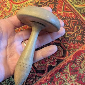 May include: A light-colored wooden mushroom-shaped object with a rounded top and a long, tapered handle. The handle has two carved lines near the bottom. The object is held in a hand against a patterned fabric background.