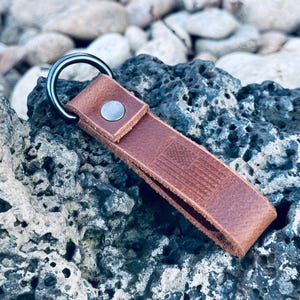 May include: A brown leather keychain with a metal ring and a silver snap. The keychain features an embossed American flag design. The leather is a medium brown color, and the keychain is resting on a dark gray rock.