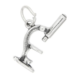 May include: A silver-colored microscope charm with a loop for attaching to a necklace or bracelet. The charm features detailed design elements, including the eyepiece, arm, and base, and is set against a white background.