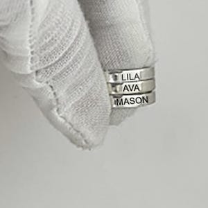 May include: Three silver rings stacked together, each engraved with a name: LILA, AVA, and MASON. The rings are held by a white fabric. The names are in a simple, sans-serif font.