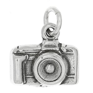 May include: A silver charm in the shape of a camera. The camera has a lens, a flash, and a body. The charm is attached to a silver loop.
