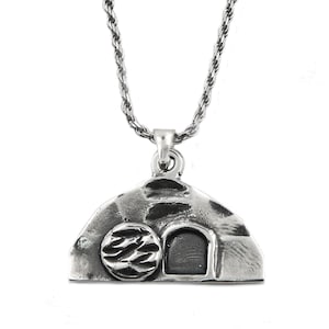May include: A silver-colored pendant necklace featuring a detailed depiction of a tomb with a stone door and a round stone. The pendant hangs from a silver rope chain. The design is detailed and textured.
