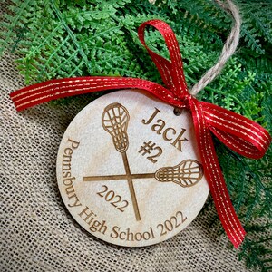 May include: A wooden lacrosse ornament with the name "Jack #2" and the text "Pennebury High School 2022". The ornament is decorated with a red ribbon with gold accents.