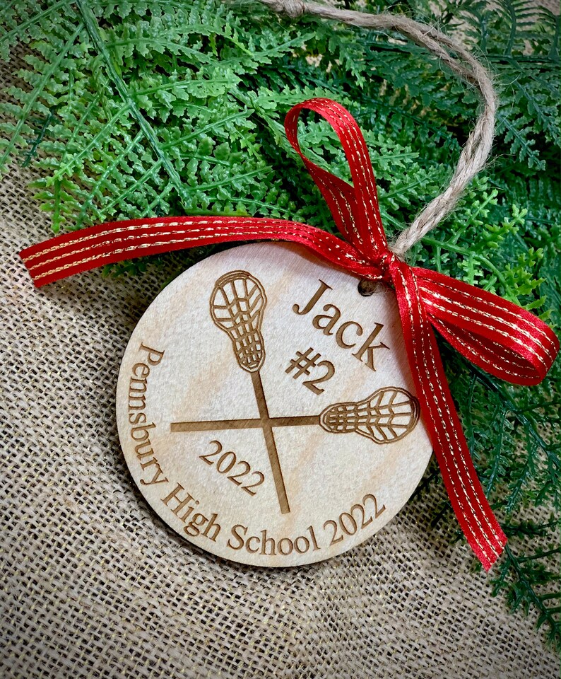 May include: A wooden lacrosse ornament with the name "Jack #2" and the text "Pennebury High School 2022". The ornament is decorated with a red ribbon with gold accents.