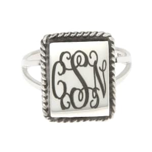 May include: A silver ring with a square-shaped white stone featuring a monogrammed design. The ring has a rope-like border around the stone.