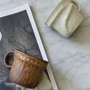 以下が含まれることがあります： ユニークなテクスチャデザインのセラミックマグカップ2つ。1つは薄いベージュ色で、もう1つは濃い茶色のアクセントが付いた茶色です。どちらのマグカップにもハンドルが付いており、白黒の画像が掲載された雑誌の上に置かれています。