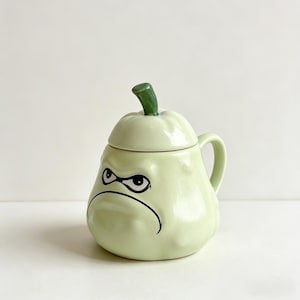 May include: A light green ceramic mug with a lid, featuring a cartoon face with a grumpy expression. The mug has a handle and a green stem-like knob on the lid. The design is a whimsical, fun style.