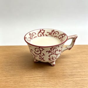 Puede incluir: Una taza de té blanca con un diseño floral rojo, llena de un líquido blanco. La taza de té tiene un asa y se apoya en tres pequeños pies. La taza está sobre una superficie de madera.