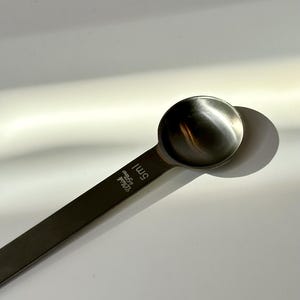 Op de afbeelding: Een roestvrijstalen maatlepel met een afgeronde kom en een lange, platte steel. De steel heeft de tekst "5ml" en de merknaam. De lepel is ontworpen voor het afmeten van ingrediënten.