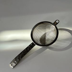 Op de afbeelding: Een roestvrijstalen thee-ei met een gehamerde handgreep. Het thee-ei heeft een cirkelvormige gaasmand en een kleine lus om op te hangen. De handgreep is donkergrijs en getextureerd, en het gaas is zilver.
