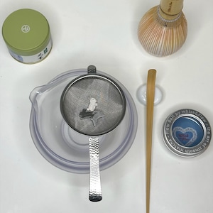 May include: A matcha tea set including a green tin, a whisk, a strainer, a bamboo scoop, a small tin with text, and a clear bowl. The strainer has a small decorative element. The set is arranged on a white surface.