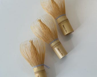 May include: Three light tan bamboo matcha whisks arranged diagonally. Each whisk features numerous thin, flexible tines and a cylindrical handle stamped with the text Whisk & Flow in a dark brown script font, secured with blue and white thread.