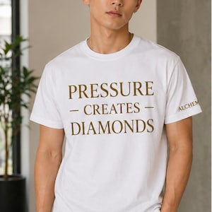 May include: White t-shirt with the phrase "PRESSURE - CREATES - DIAMONDS" printed in brown. The word "ALCHEM" is printed on the right sleeve. The shirt is worn by a person.