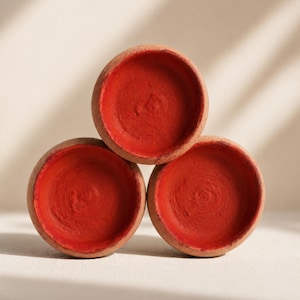 Op de afbeelding: Drie kleine, ronde terracotta kommen op elkaar gestapeld. De binnenkant van elke kom is felrood geschilderd, wat contrasteert met de natuurlijke, ongeverfde buitenkant. De kommen zijn in een driehoekige formatie gerangschikt.
