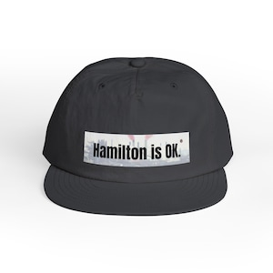 May include: A black baseball cap with a curved brim. The cap features the text "Hamilton is OK." in a white rectangular patch. The text is in a bold, sans-serif font. The background of the patch appears to be a blurred image.