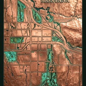 May include: A copper-colored relief map of Bozeman, Montana, with the city name at the top. The map features detailed streets and waterways, with green accents. The map is framed in a dark green border.