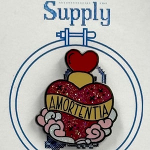 May include: A pin with a heart-shaped design, featuring the word "Amortentia" in gold lettering. The pin has a red, yellow, and pink color scheme, with glitter accents. The text "27 Stitch Supply" is at the top.