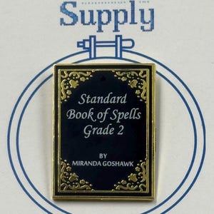 May include: A rectangular needle minder with a black background and gold trim, featuring the text "Standard Book of Spells Grade 2" and the author's name. The design is set against a blue and white background with the text "27 Stitch Supply" at the top.