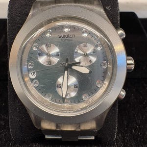 May include: A Swatch Swiss chronograph watch with a dark grey dial, three silver sub-dials, and clear crystal hour markers. The watch features a translucent grey plastic case, a scratched watch face, and white-tipped hour and minute hands.