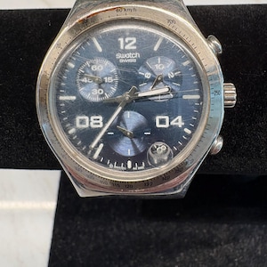 May include: A Swatch Swiss chronograph watch with a dark blue dial, silver-tone hands, and large white numerals at 12, 08, and 04. The watch features a tachymeter scale on the silver bezel, three sub-dials, and a date window at the 4 o'clock position.