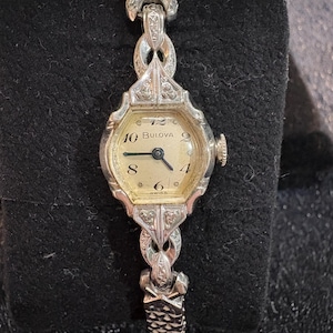 May include: A vintage Bulova wristwatch with a silver-toned metal strap and a cream-coloured face. The watch face features black numerals and hands, with the brand name "Bulova". An elegant accessory.