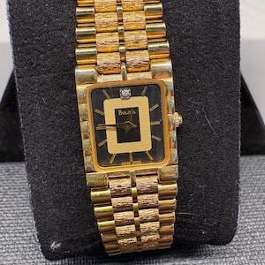 May include: A gold-tone Bulova wristwatch featuring a rectangular black dial with a central gold square, a single diamond marker at the twelve o'clock position, and a textured link bracelet with a polished finish and gold-tone metal construction.