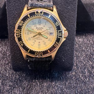 Pode incluir: Um relógio Elgin dourado com moldura preta e pulseira de couro preta. O mostrador do relógio é dourado claro com algarismos e ponteiros pretos. O relógio exibe a palavra "ELGIN" e "QUARTZ".