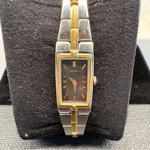 May include: A Seiko wristwatch featuring a rectangular black dial with gold-tone hour markers and hands. The watch has a two-tone metal link bracelet in silver and gold. The case is rectangular with a gold-tone bezel and a crown on the right side.