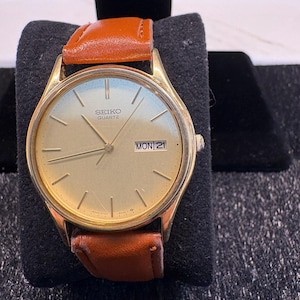 May include: A gold-tone Seiko Quartz wristwatch featuring a round champagne-colored dial with thin gold hour markers, gold hands, and a day-date window at the three o'clock position. The watch is fitted with a smooth, tan-colored leather strap.