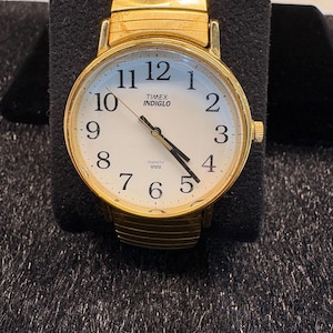 Könnte beinhalten: Eine goldfarbene Timex Indiglo Quarzuhr mit weißem Zifferblatt und schwarzen Ziffern. Die Uhr hat ein goldenes Dehnband und wird auf einer schwarzen Oberfläche präsentiert.
