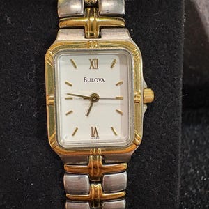 May include: A Bulova wristwatch with a rectangular white face, gold-tone bezel, and silver and gold-tone band. The watch face displays Roman numerals and the Bulova brand name. The watch has a gold crown and a silver and gold-tone band.
