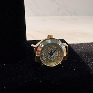 May include: A gold-toned ring watch with a round face featuring an eagle emblem and the words "UNITED STATES OF AMERICA". The watch has a black band and a gold crown. The watch is displayed on a black velvet surface.