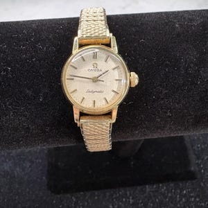 May include: A gold-toned Omega Ladymatic wristwatch with a textured band and a round face. The watch features a gold bezel, hands, and hour markers. The watch face is cream-colored with the Omega logo and the word "Ladymatic" printed on it.
