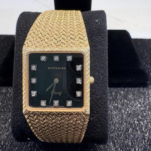 Könnte beinhalten: Goldfarbene Wittnauer Quartz-Uhr mit quadratischem, schwarzem Zifferblatt. Die Uhr hat Diamant-Stundenmarkierungen und ein gewebtes Goldarmband. Die Uhr wird auf schwarzem Samt präsentiert.
