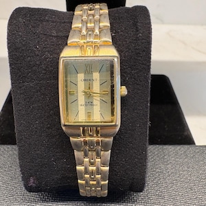 May include: A rectangular Orient wristwatch with a two-tone gold and silver metal link band. The watch face features a vertical striped pattern, gold-tone hands, a Roman numeral at the twelve o'clock position, and text reading Orient, 3 ATM WATER PROOF.