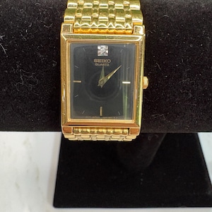 May include: A gold-toned Seiko Quartz watch with a rectangular black face. The watch features a gold band and a small diamond accent near the top of the face. The watch is displayed on a black surface.