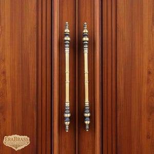 May include: Two brass door pulls with a bamboo-like design are mounted on a wooden door. The pulls have a gold finish with dark blue accents. The door is made of wood with vertical grain patterns. The logo "ERABRASS ANTIQUE HARDWARE" is visible.