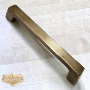 May include: A rectangular brass cabinet pull with a square handle design. The antique hardware has a warm, golden-brown color and is set against a white wood background. The logo "ERABRASS ANTIQUE HARDWARE" is visible.