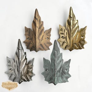 May include: Four metal leaf-shaped decorative hardware pieces in bronze, gold, silver, and verdigris finishes. Each leaf features detailed veining and two mounting holes. The EraBrass Antique Hardware logo is visible in the lower left corner.