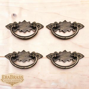 Puede incluir: Cuatro tiradores de cajón de latón antiguo con un diseño decorativo de hojas. Cada tirador presenta un asa curvada y una placa trasera detallada. Los tiradores están dispuestos sobre una superficie de madera clara. La marca "ERABRASS ANTIQUE HARDWARE" es visible.