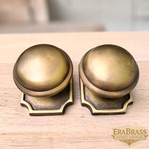 May include: Two round brass door knobs with a smooth, domed surface and a dark, aged patina. Each knob is mounted on a square-shaped backplate with rounded corners and decorative edges. The text "ERABRASS ANTIQUE HARDWARE" is visible in the corner.