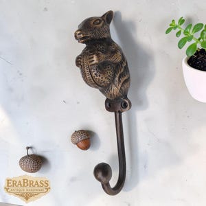 May include: A bronze squirrel-shaped wall hook with a curved hook. The squirrel is holding an acorn. Two acorn-shaped decorative objects are also present. The hook is mounted on a light-coloured wall. The logo "ERA BRASS ANTIQUE HARDWARE" is visible.