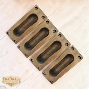 Puede incluir: Cuatro tiradores empotrados de latón antiguo. Cada tirador rectangular tiene una hendidura ovalada central para agarrar y cuatro orificios para tornillos. Los tiradores están dispuestos diagonalmente sobre una superficie de madera clara. El logotipo "ERABRASS ANTIQUE HARDWARE" es visible.