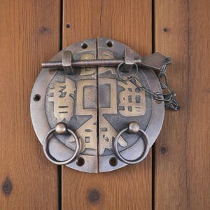 May include: A bronze-colored, circular door latch with Chinese characters and two ring pulls. The latch is mounted on a wooden door with vertical planks. A metal chain and key-shaped piece are attached.