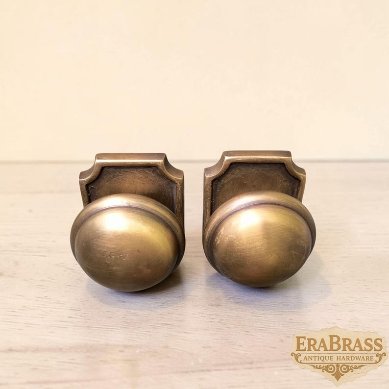 May include: Two matching brass door knobs with a rounded, smooth front and a rectangular backplate featuring a curved top edge. The metal has a brushed, antique finish with visible golden tones. The text EraBrass Antique Hardware is visible.